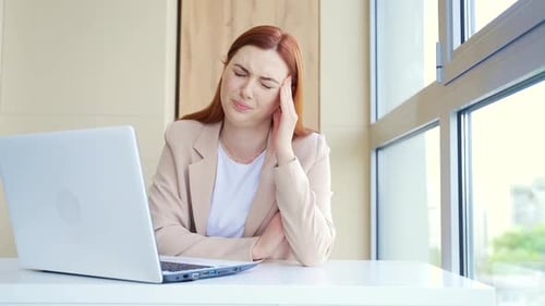 Close-up portrait of a young red-haired woman suffering from a severe headache at work in workplace.
