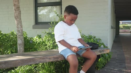 Afroamerikanisches Kind konzentriert sich auf das Tablet, sitzt draußen auf einer Bank und klopft, umarmt sich und lächelt im Tee