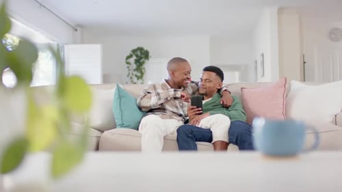 Happy african american gay male couple sitting on sofa, embracing and using smartphone, slow motion