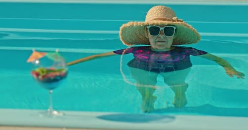 Sweet Elderly Woman Cooling Off in a Pool with a Cold Drink with Ice on the Pool Edge