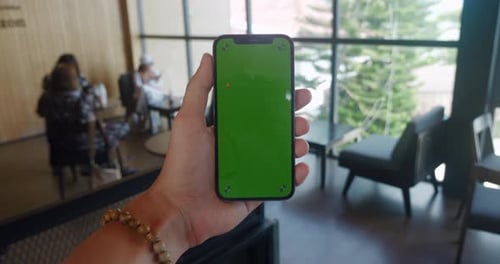 Male Hand Holding Smartphone with Green Screen Chroma Key on the Background of Cafe Filled with