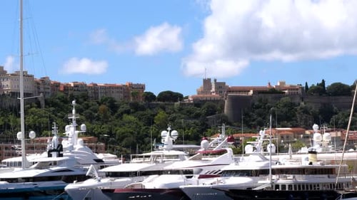 Wide shot of luxury yachts at marina and Prince Palace Building on hilltop. Sunny day with puffy clo
