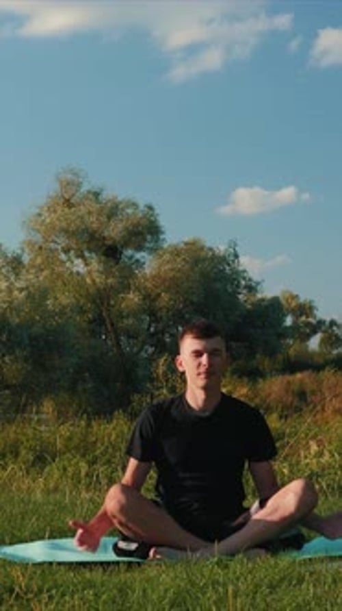 Young adult sportsman meditating in lotus pose on yoga rug at city park. Vertical video.