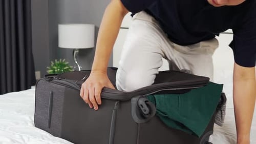 man trying to close overfilled suitcase on his bed