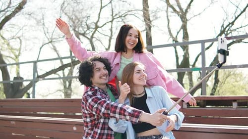 Three Friends Taking Selfies in a Sunny Park with Selfie Stick Capturing Joyful Moments Together