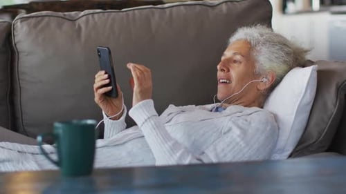 Senior mixed race woman lying on sofa wearing earphones and using smartphone