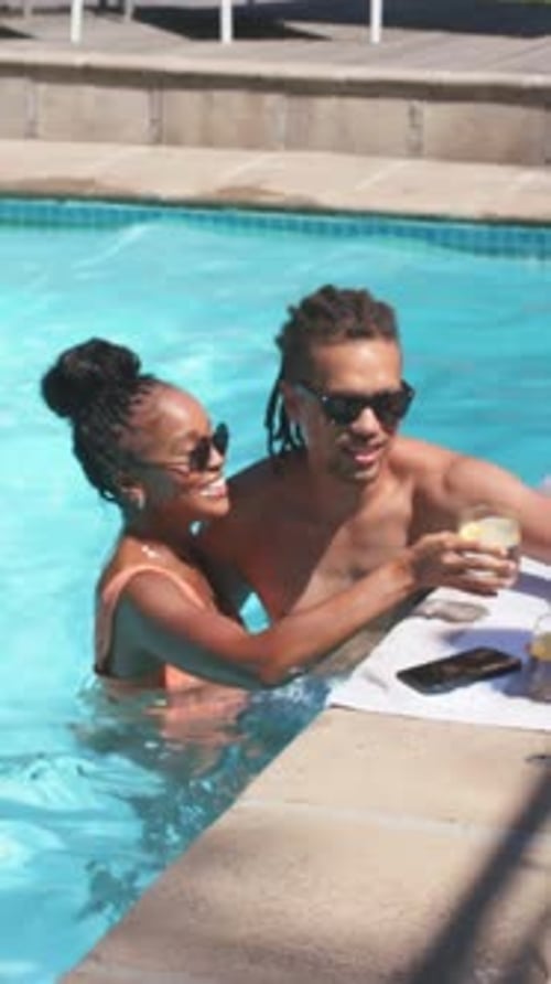 Vertical video: Hugging woman in swimsuit reaching for drink on edge, giving glass to man at pool