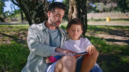 Cute Family Reading Book Sitting Grass at Forest Closeup Father Son Under Tree