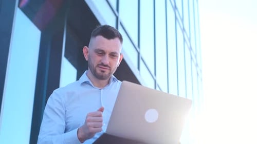 A Business Man Works on a Laptop Near the Office Center Against the Background of the Sun's Rays