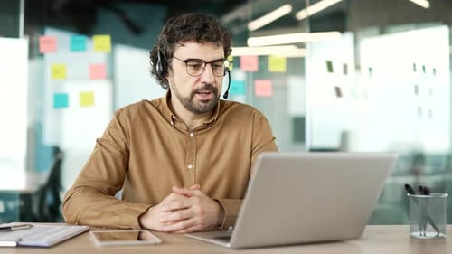 Confident businessman in wireless headset talking on a video call using laptop sitting at workplace