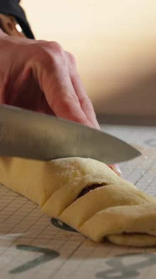 Close-up Of Hand Using Kitchen Knife In Slicing Log Of Rolled Dough For Cinnamon Rolls. vertical