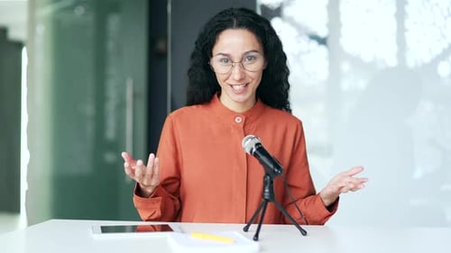 Webcam view. Young female employee with a microphone is recording a podcast sitting at workplace