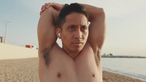 Close up, young athletic man standing on the beach stretching his arm muscles before training