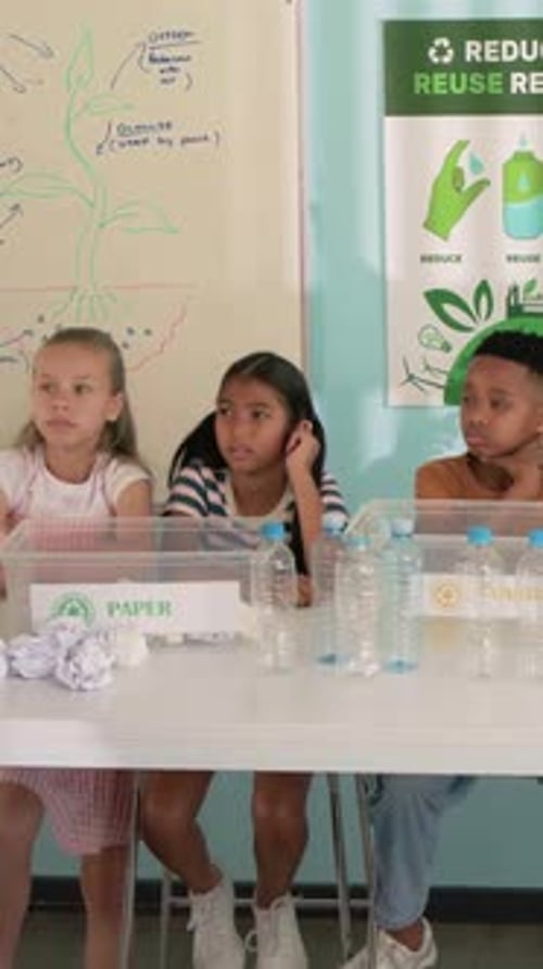 Vertical video: Talking center student engaging peers near PAPER bin during recycle class at table