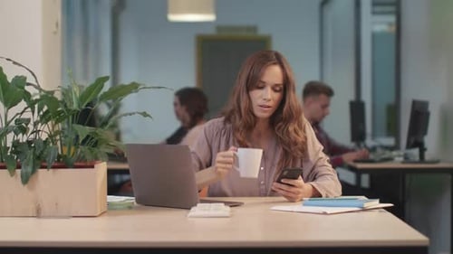 Woman Drinks Coffee While Using Phone in Office