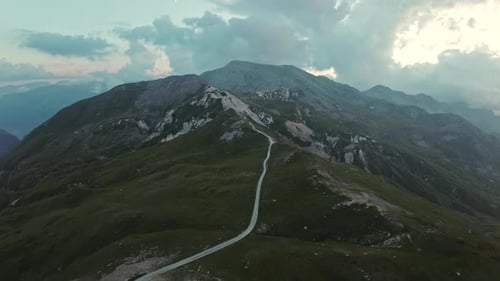 A winding road stretches over green mountain peaks under a cloudy sky at sunset