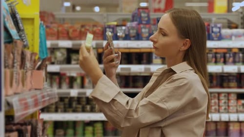 A Woman in a Grocery Store Aisle Reaches for a Can of Cat Food Her Smartphone in Hand She is About