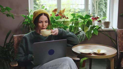 Woman Enjoying Coffee in a Cozy Cafe While Working on a Laptop Surrounded By Plants and Natural