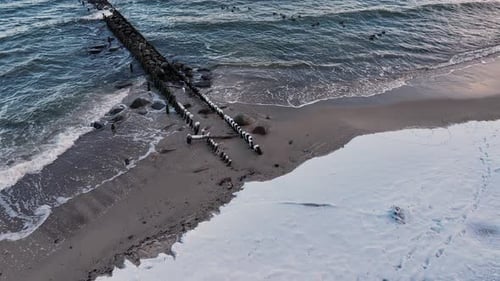 Waves Crash Against Sea Snow Covered Breakwater Along Sandy Beach in Late Afternoon Light Near