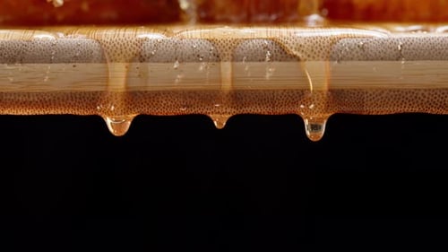 Drops of honey dripping from a wooden board onto a black background, close-up with a camera tilt.