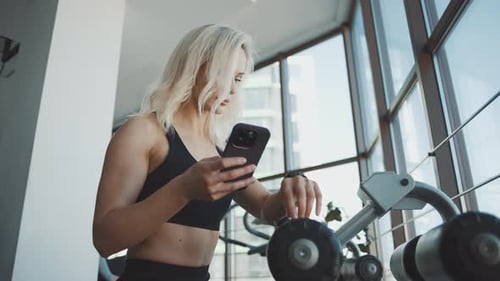 Girl checking phone in gym while preparing to lift weights in a bright workout space
