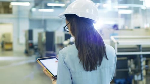 Beautiful Female Industrial Engineer in a Hard Hat Uses Tablet Computer while Walking in Big Factor