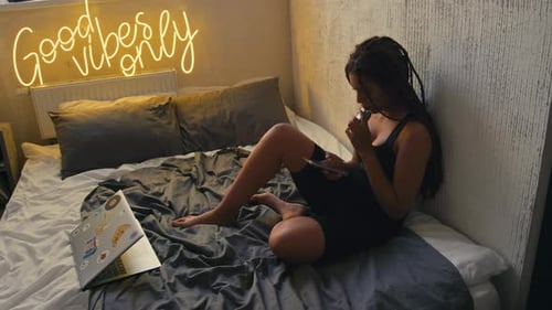 Chica vapeando sentada en la cama en una habitación tipo loft con letrero de neón Good Vibes Only