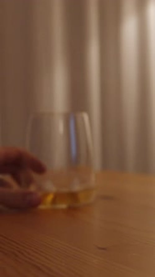 Vertical video. A glass of whiskey on a wooden table. A man brings it closer to the camera.