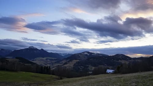 Eine hügelige ländliche Landschaft, Wolken, die sich in der Abenddämmerung bewegen, Graswiesen mit Bäumen, Bergen