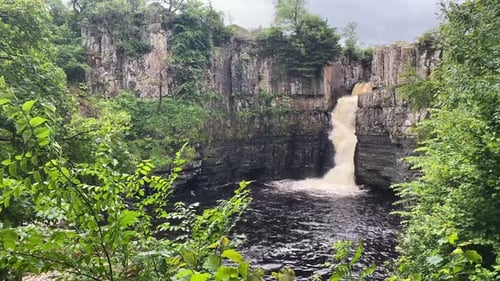 High Force Waterfall through some green trees in a cloudy day. Teesdale, North-East England UK, Stil