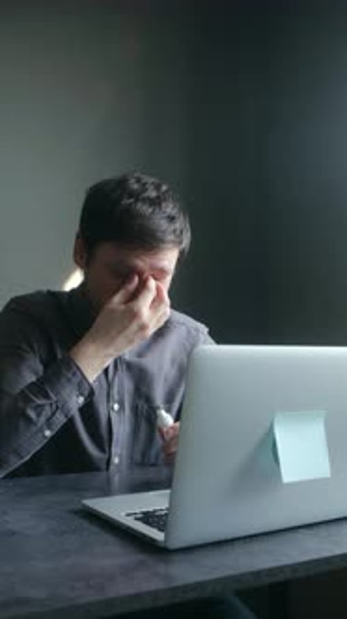 Müder Mann reibt sich die Augen, während er zu Hause an einem Laptop arbeitet