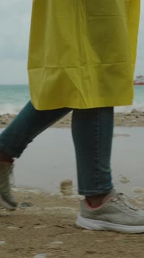 A view of a woman's legs in jeans and sneakers, she walks on the seaside beach in a yellow raincoat
