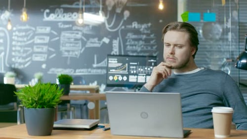 Handsome Young Office Employee Thinks on a Problem Solution While Working on a Laptop Computer. He'