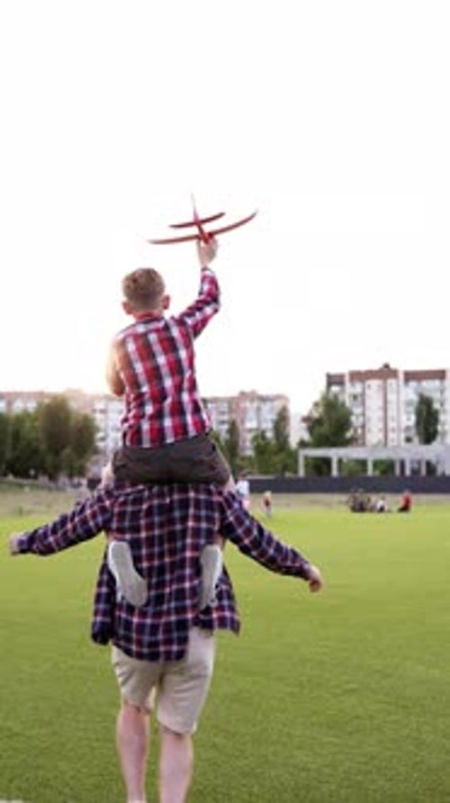 Father carrying child on shoulders with airplane in hand at sunset in park. Happy family dreams.