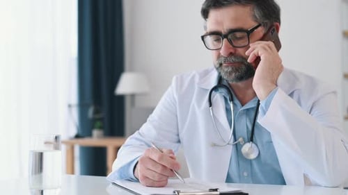 Talking by phone and writing data. Mature male doctor in coat is in the clinic room