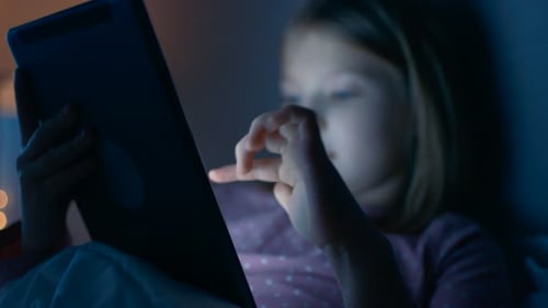 Une jolie petite fille la nuit dans son lit interagit avec une tablette informatique.