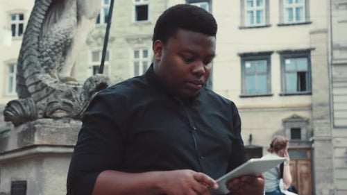Handsome Serious AfroAmerican Man in Elegant Outfit Sits By the Monument in the Central Square Surfs