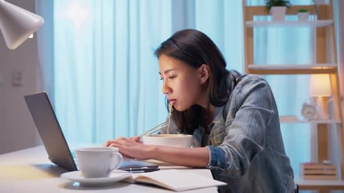 Young Adult Eats Noodles While Working on Laptop