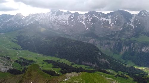 Drone shot (rotating) of a Mountain Summit covered in grass with a rocky Mountain Range in the Backg