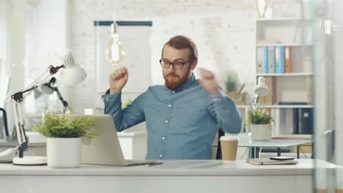 Un technicien barbu commence à danser alors qu'il est assis à son bureau dans un bureau bien éclairé.