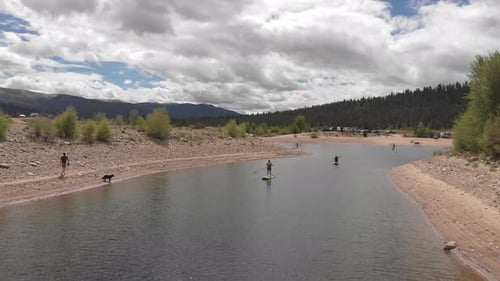 stand up paddle boarding in a channel of a lake in the mountains while a dog runs down the beach