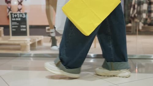 Füße laufen mit gelber Tasche über glänzende Fliesen im Einkaufszentrum, Jeans und Sneakers in Bewegung, Lowangle Shot