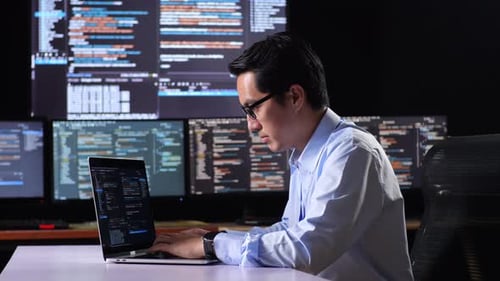 Side View Of Asian Male Programmer Writing Code By A Laptop Using Multiple Monitors