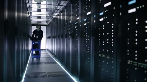 Un ingénieur informatique pousse un chariot de secours dans le couloir du centre de données avec des rangées de serveurs rack. Il ouvre un rack