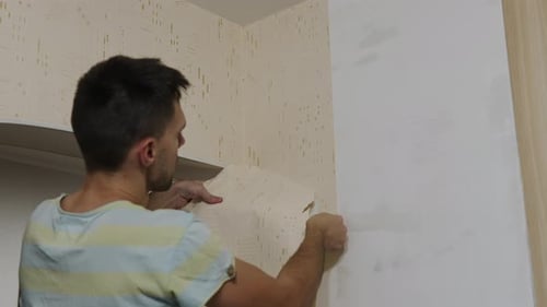 Kaukasischer Mann in der Wohnung reißt ein Stück Tapete von der Wand im Innenbereich