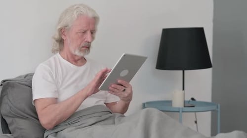Senior Man Uses Tablet in Bed At Home