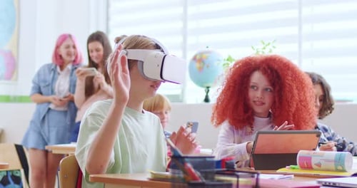 Little Caucasian Boy Pupil in VR Headset Sitting at Desk in Classroom and Having Virtual Reality
