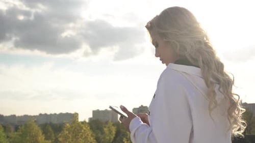 A Woman Writes a Message on a Smartphone Against the Backdrop of the City and Sunset