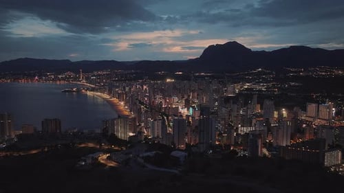Waterfront city with bright lights next to calm blue ocean at night.