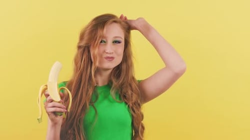 Cheerful Young Woman Dances in Front of the Camera and Eats a Banana A Redhaired Woman in a Green